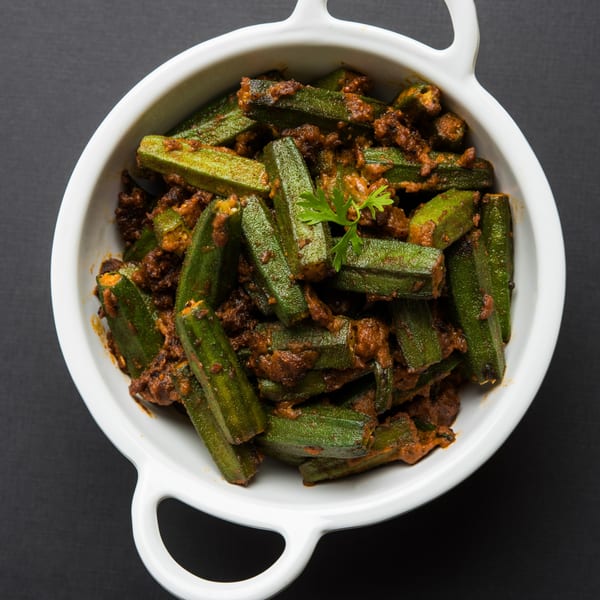 Spicy okra curry, also known as Bhindi Masala, in a white bowl with a sprig of cilantro.