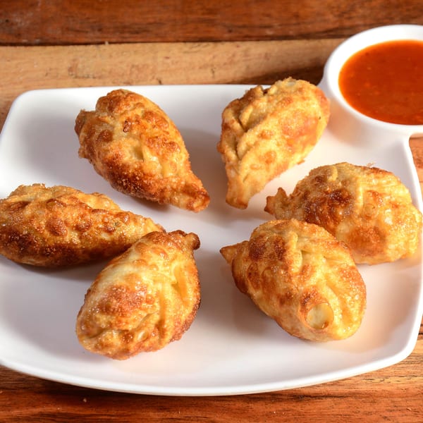 Six golden-brown fried dumplings arranged on a white plate with a side of red dipping sauce.