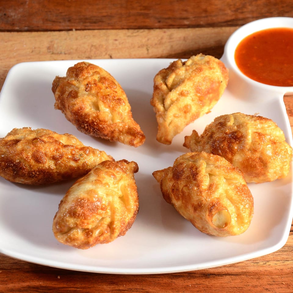 Six golden-brown fried dumplings arranged on a white plate with a side of red dipping sauce.