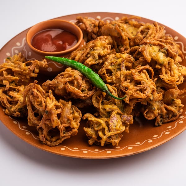 Crispy onion pakoras with a side of red dipping sauce and a green chili on a brown plate.