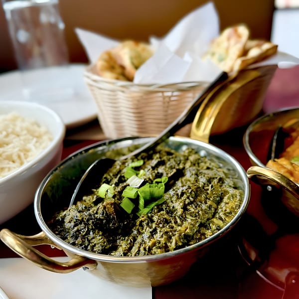 A brass bowl of green saag, topped with scallions, next to rice and naan bread.